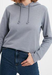 Sudadera gris claro con un bolsillo frontal, cordones ajustables y mangas largas, combinada con jeans de mezclilla azul de cintura alta.