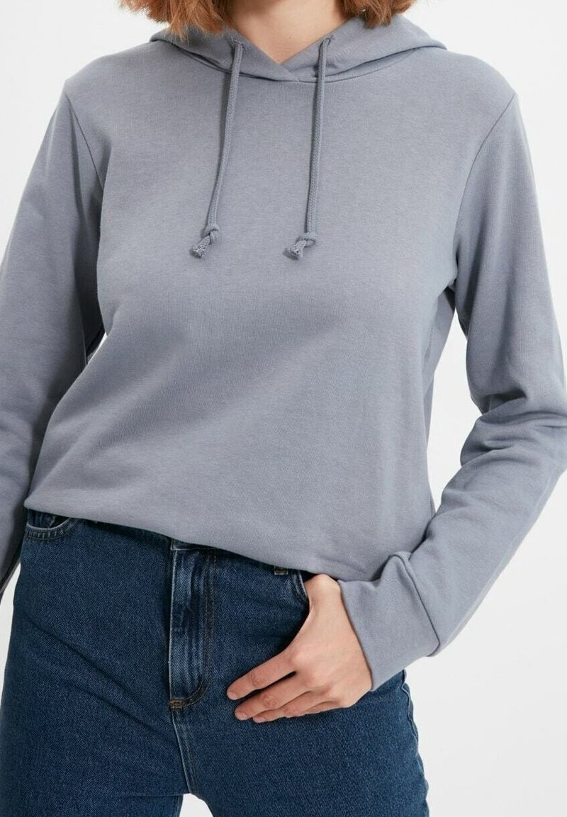 Sudadera gris claro con un bolsillo frontal, cordones ajustables y mangas largas, combinada con jeans de mezclilla azul de cintura alta.