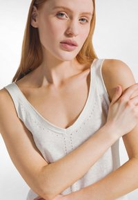 Canotta bianca con scollatura a V profonda, realizzata in materiale leggero con piccole perforazioni lungo la parte centrale anteriore. Texture liscia.