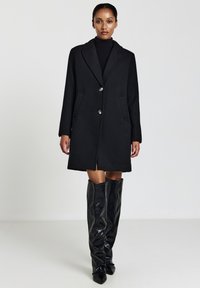 Donna che indossa un cappotto nero lungo fino al ginocchio con bottoni, un dolcevita nero e stivali neri lucidi fino al ginocchio, in piedi davanti a uno sfondo neutro.