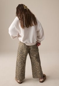 Sweat-shirt blanc surdimensionné à épaules tombantes, associé à un pantalon large imprimé léopard beige et marron, complété par des chaussures marron glissantes.