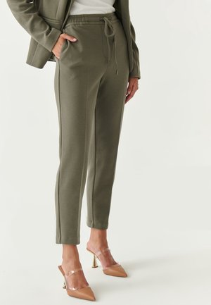 Žena nosící olivově zelené kalhoty se stahovacími šňůrkami, béžové vysoké pantofle s průhlednými pásky a ladící olivový blejzr přes bílé tričko.