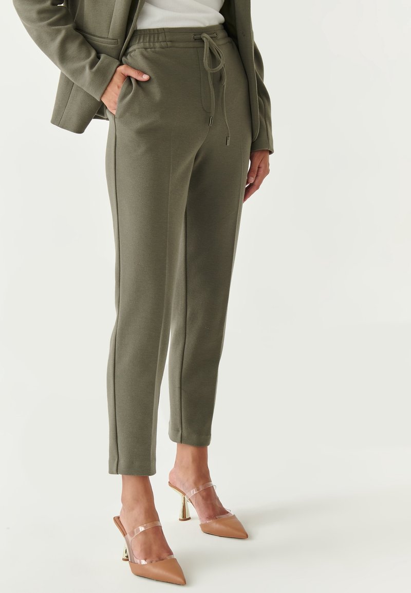 Femme portant un pantalon à cordon de serrage vert olive, des mules à talons hauts beiges avec des brides transparentes, et un blazer assorti vert olive sur un haut blanc.