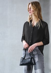 Bolso de piel negra con asas cortas, textura suave y forma triangular, sostenido por una persona que lleva una camisa negra transparente y jeans grises.
