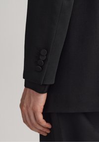 Manica di giacca nera con texture liscia, quattro bottoni rivestiti in tessuto e vestibilità su misura. Nessun motivo visibile.