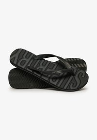 Sorte flip-flops med teksturerede såler, der har et fremtrædende fiskebensmønster og hævet "SUPERDRY" branding hen over fodsengen.