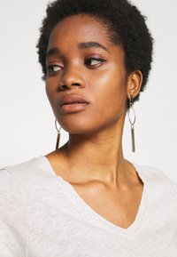 Gaiši pelēks krekls ar V veida kakla izgriezumu un strukturētu audumu, apvienots ar zelta toņu auskariem ar karājošiem elementiem, kas ir ovālas formas ar vertikālām svītrām.