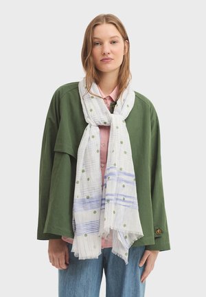 Veste verte surdimensionnée avec une finition texturée, portée par-dessus une chemise rose clair, associée à une écharpe blanche ornée de points verts et de rayures bleues.