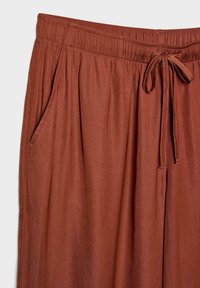 Roestkleurige, lichte broek met een elastische tailleband, koord en zijzakken. Gemaakt van zachte stof met een gladde textuur.