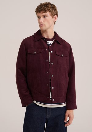 Jeune homme portant une veste boutonnée rouge foncé par-dessus une chemise rayée et un jean bleu foncé, regardant sur le côté sur un fond uni.