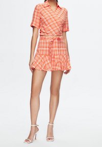 Tvådelat set som består av en kortärmad, insvängd toppskjorta och en svängd minikjol i orange rutigt mönster. Materialet har en slät textur med volangdetaljer.