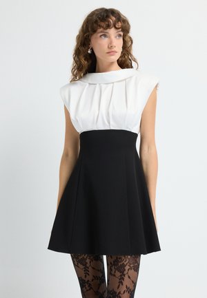 Jurk met een witte geplooide lijfje en korte mouwen, overgaand in een nauwsluitende zwarte rok. Gecombineerd met zwarte gemusterde panty’s.