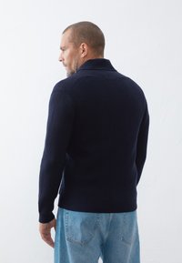 Navy blue knitted sweater with a high collar, ribbed texture, and a fitted cut, paired with light blue denim jeans.