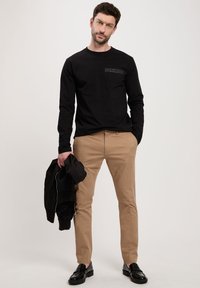 Czarna koszula z długim rękawem z kieszonką na piersi, beżowe spodnie slim fit i czarne buty; trzyma czarną kurtkę; jednolite tło.