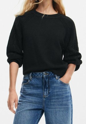 Pull en tricot noir avec des manches en tricot torsadé, un col rond et des poignets côtelés. Associé à un jean bleu taille haute.