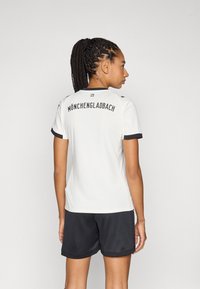 Bijela sportska majica s crnim naglascima, s natpisom "MÖNCHENGLADBACH" na leđima, kratke rukave i u kombinaciji s crnim kratkim hlačama.