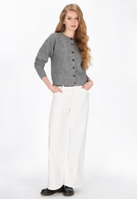 Grau gestrickte Strickjacke mit einem strukturierten Rautenmuster und braunen Knöpfen, kombiniert mit weißen Hose mit weitem Bein und schwarzen Stiefeletten.