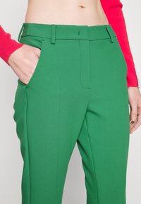 Pantalons tailleur verts en tissu lisse et texturé. Les caractéristiques incluent une coupe droite, des poches latérales et une taille plate.