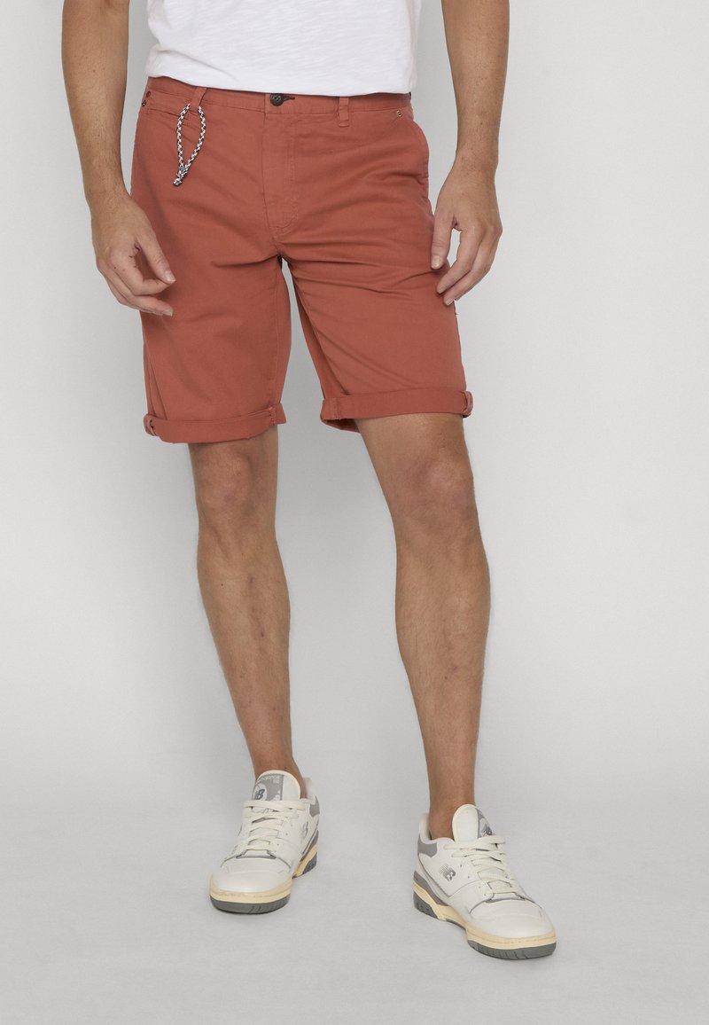 Homme portant un short rouille retroussé, des baskets blanches et un t-shirt blanc, debout devant un fond clair uni.