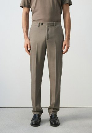 Hombre vistiendo pantalones a medida color topo, camisa de manga corta a juego color topo y mocasines de cuero negro, de pie contra un fondo blanco liso.