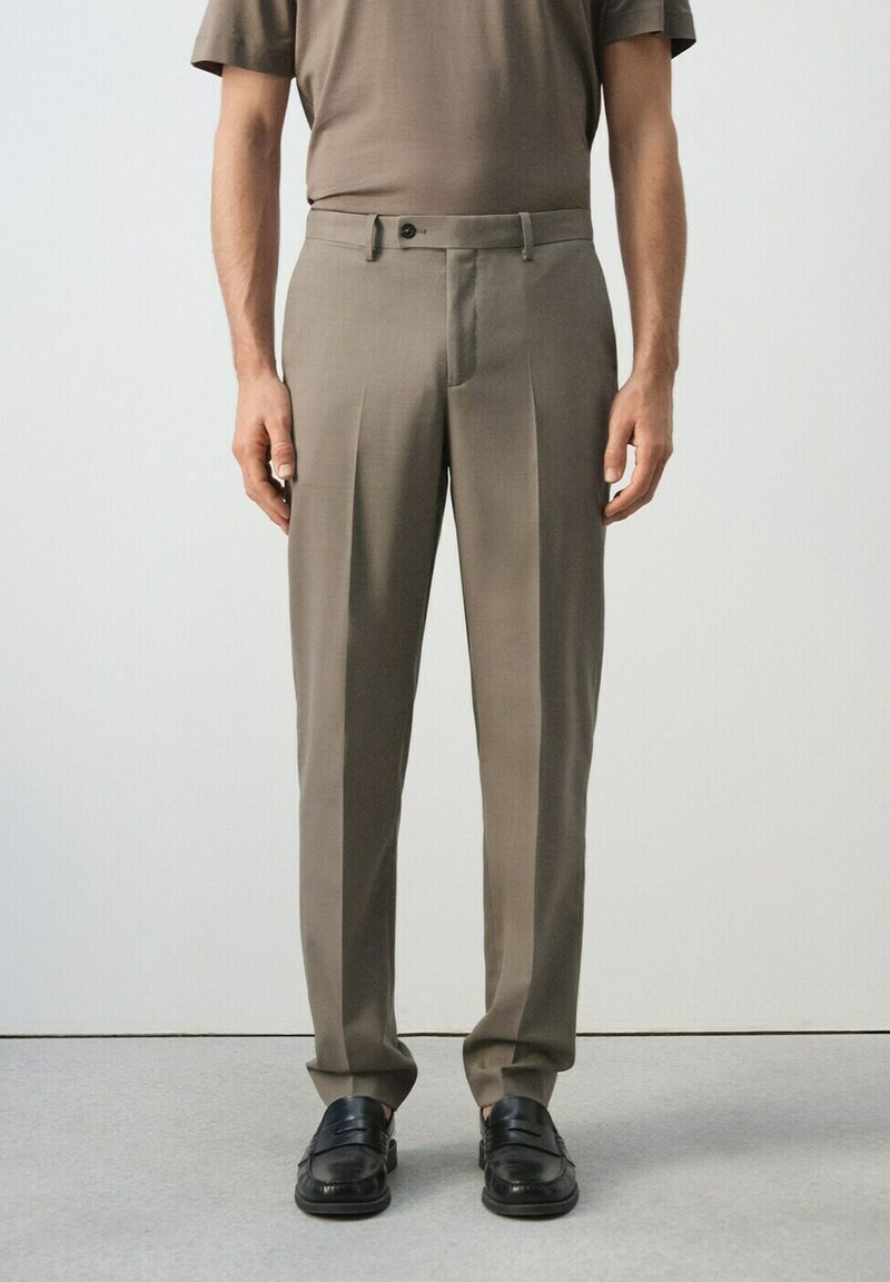 Muž oblečený v taupe šitých nohaviciach, zodpovedajúcej taupe košeli s krátkym rukávom a čiernych kožených mokasínach, stojaci na bielom pozadí.