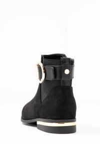 Botín negro con una mezcla de ante y tela suave, acento en forma de hebilla redonda dorada, ribete dorado en la suela y un tacón negro sólido.