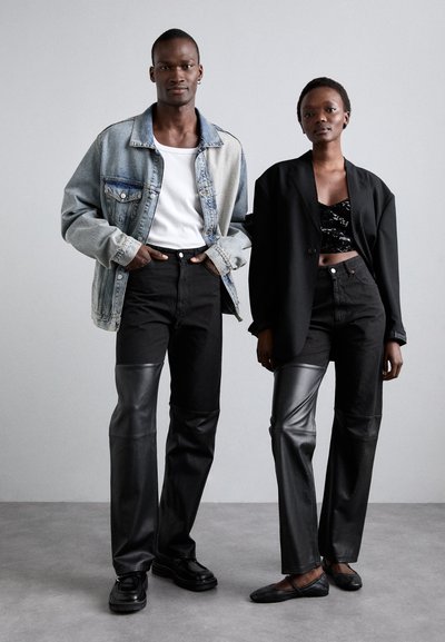 Two models stand side by side. The man wears a denim jacket, white shirt, black jeans; the woman has a black blazer, sequined top, and black pants with leather accents.