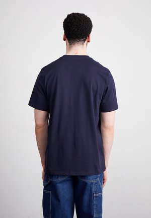 Mand med kort krøllet hår står med ryggen til iført en ensfarvet mørkeblå t-shirt og blå denim cargobukser mod en ensfarvet lys baggrund.