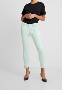 Mint green skinny jeans with a five-pocket design, button closure, and a smooth texture, paired with a black short-sleeve t-shirt and black heels.