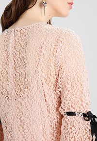 Gros plan d'une femme portant un haut en dentelle rose pâle avec un ruban noir noué sur la manche et une boucle d'oreille florale décorative.