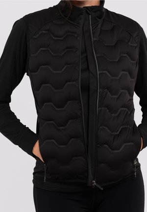 Kobieta ubrana w czarną pikowaną kamizelkę bez rękawów na koszulę z długim rękawem, z rękami w kieszeniach kamizelki, przedni zamek częściowo otwarty.