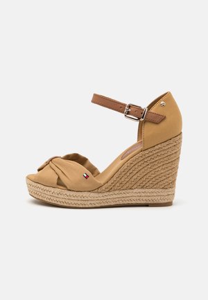 Tan kile-sandal med flettet platform, ankelrem med spænde og frontknude med et lille emblem i rød, hvid og blå.