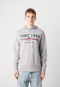 Hoodie gris avec une poche kangourou, une capuche à cordon de serrage et un logo imprimé avec des accents rouge et marine. Porté avec un jean bleu clair usé.