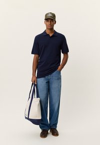 Μπλε ναυτικό πόλο πουκάμισο, ανοιχτό μπλε τζιν με φαρδιά πόδια, καφέ παπούτσια, ελιά καπέλο με γραφικό και λευκή πάνινη τσάντα με μπλε ναυτικές λεπτομέρειες.