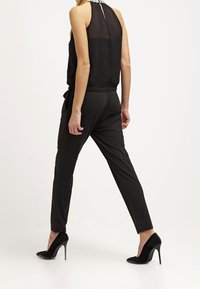 Mujer caminando, vistiendo una blusa sin mangas de color negro con detalles en el cuello blanco, pantalones negros ajustados y zapatos de tacón negro, mostrada de espaldas.