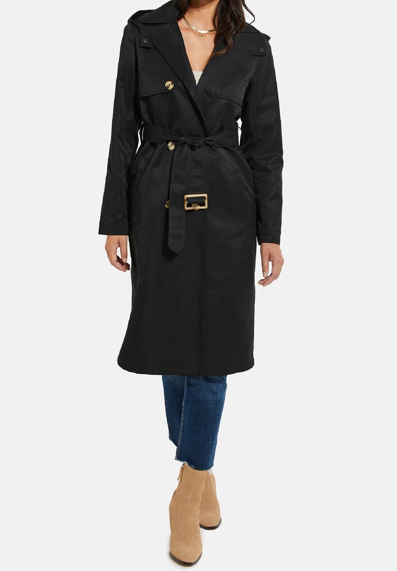 Schwarzer Trenchcoat mit einem taillierten Gürtel, goldenen Beschlägen, doppelreihigen Knöpfen und einem klassischen Kragen. Getragen über blauen Jeans und Ankle Boots.
