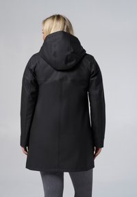 Veste noire imperméable avec capuche, dotée d'une texture lisse, de manches longues et de détails de couture visibles. Portée avec un pantalon gris.