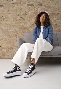 Svarta högskurna sneakers med vit sula, snörning och sidologotyp. Bärs med vita vida byxor och en ljus denimskjorta, sittande på en grå soffa.