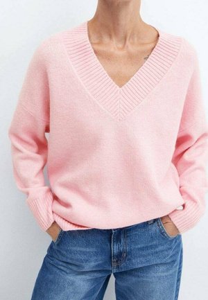 Person iført en lyserød V-hals sweater med ribkanter og blå denim jeans, hænderne stikket i forlommerne, stående mod en hvid baggrund.