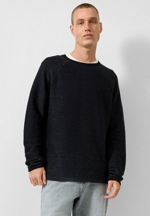 Jonge man met kort haar die een zwarte gestructureerde trui draagt over een wit overhemd en lichtgrijze spijkerbroek, staand tegen een effen achtergrond.