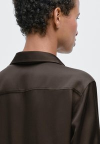 Camicia marrone in raso con colletto, caratterizzata da una texture liscia e una leggera lucentezza. Sul retro presenta una cucitura a yoke sulle spalle.