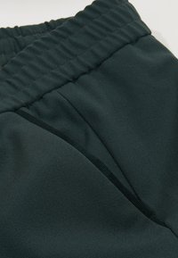 Tmavě zelený materiál s texturovaným povrchem a elastickým pasem, se šitými švy a jemnými záhyby pro uvolněný střih.