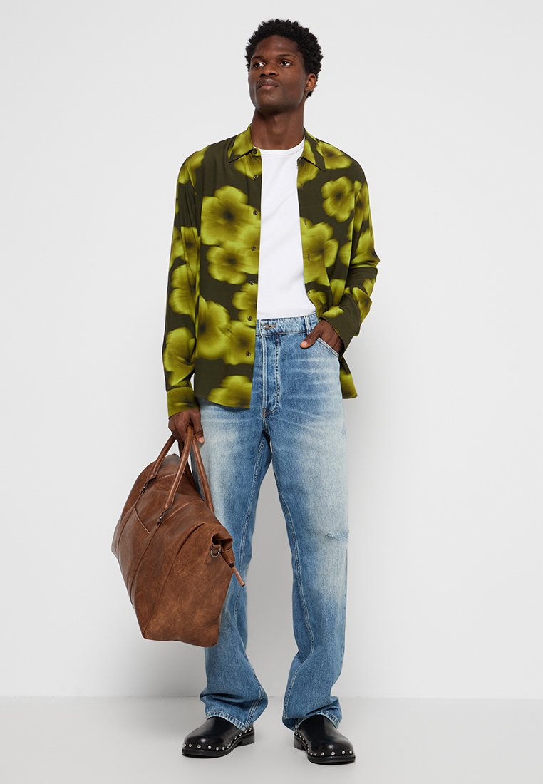 Homme portant une chemise à boutons verte à fleurs, un t-shirt blanc, un jean bleu clair, des chaussures noires à clous, et tenant un grand sac en cuir marron.