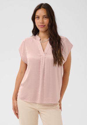 Femme aux longs cheveux foncés portant un chemisier rose pâle à manches courtes et un pantalon beige, debout devant un fond clair uni.