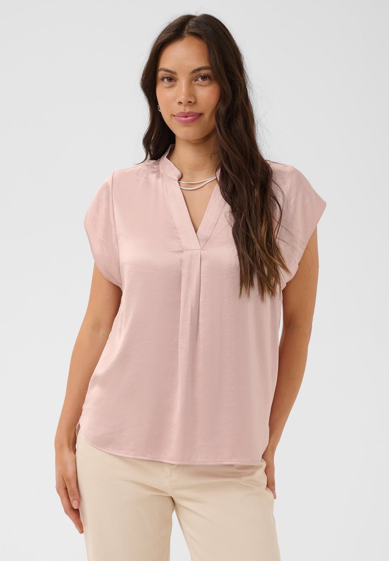 Femme aux longs cheveux foncés portant un chemisier rose pâle à manches courtes et un pantalon beige, debout devant un fond clair uni.