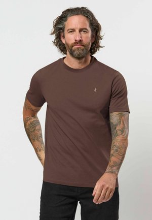 Mand med krøllet hår og skæg iført brun t-shirt og sorte bukser, der viser detaljerede tatoveringer på begge arme mod en ensfarvet baggrund.