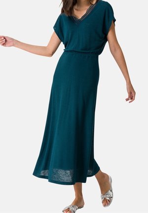 Frau trägt ein tealfarbenes Midi-Kleid mit V-Ausschnitt und Spitzenbesatz sowie silberne Slide-Sandalen und posiert mit ausgestreckten Armen vor einem weißen Hintergrund.