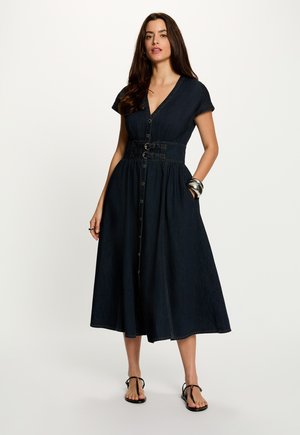 Mujer con cabello largo y oscuro, vistiendo un vestido midi de mezclilla oscuro abotonado con detalle de cinturón, sandalias negras y pulseras plateadas sobre un fondo blanco.
