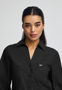 Svart skjorta med knappar och krage, med två bröstfickor och en liten logomärke. Slät textur och klassisk skärning.