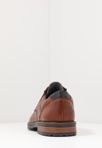 Zapato de cuero marrón con un acabado liso, punta redondeada y acento en el tacón negro. Suela de goma de bajo perfil con un dibujo texturizado para mayor agarre.